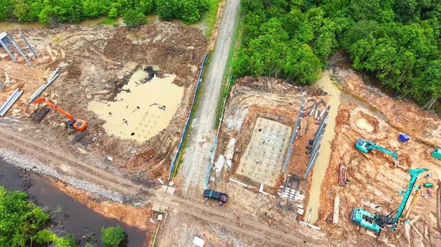 Aerial drone orbiting shot over a bridge construction site showing concrete foundation piles, excavator, and road infrastructure development in a green environment.
