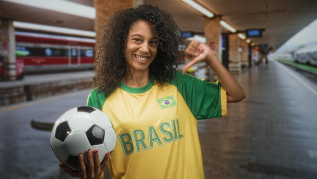 Woman holding a soccer ball and points thumb to chest wearing a brasil team t shirt in a train station building; pride support fandom.