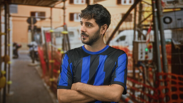 Young hispanic man with beard in blue football shirt and team jersey, arms crossed and pouting while standing by construction scaffolding on a street; disappointment.