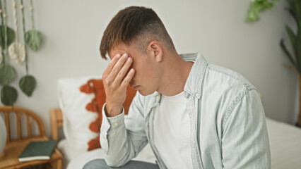 Obraz premium Man pressing forehead with hand while sitting on bed beside pillow and book in a building; fatigue headache.