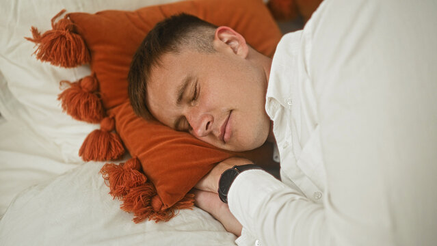 Man sleeping on side with face on orange tassel pillow on bed in bedroom, wearing white shirt and wristwatch visible; serenity.