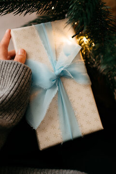 A person wearing a cozy grey sweater holds a gift box wrapped in star-patterned paper with a light blue frayed-edge ribbon, set against a dark Christmas tree background illuminated by warm, glowing fa