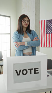 Young woman with arms crossed wearing an i voted sticker at a voting booth in a public building with an american flag; pride civic duty.
