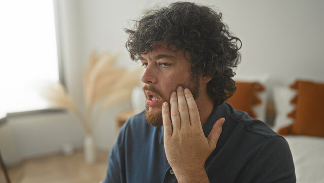 Young hispanic man with curly hair and beard holding cheek with hand, wincing on bed inside a building; toothache pain.