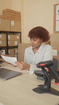 Woman claps hands and holds documents at a desk with cardboard boxes and a barcode scanner in an office building; small business success joyful.