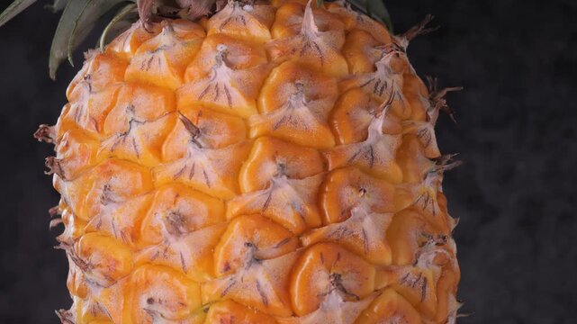 Close-up rotating shot of pineapple skin texture with leaves on a dark background.	