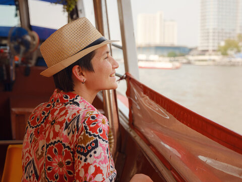 Female tourist in elegant summer dress and straw hat enjoys scenic boat ride along Chao Phraya River in Bangkok. Woman looks at waterfront view and enjoys refreshing breeze.