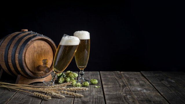 Rustic beer still life with wooden barrel and glasses, fresh craft beer with foam on dark background, brewery concept with hops and wheat