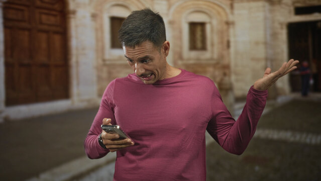 Man with moustache scrolling a smartphone with hands visible while shrugging one hand on a cobbled street by a stone building and wooden door; mild confusion.