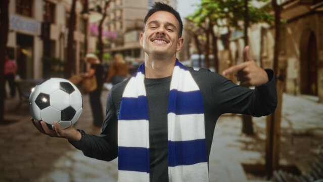 Hispanic man with moustache wearing blue and white scarf holding soccer ball and points finger to ball in cobblestone street; pride team spirit.