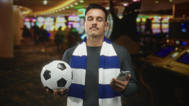 Man with mustache wearing a blue and white scarf, holding a soccerball in one hand and a smartphone in the other inside a casino while checking his phone; anticipation camaraderie.
