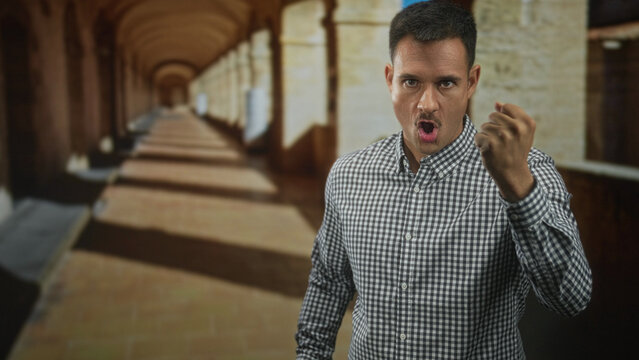 Young hispanic man with moustache clenches fist forward in arched building corridor wearing check shirt; anger defiance tension.