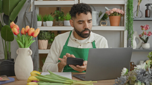 Man florist in green apron at counter with tulip vase holds card reader and laptop, hand to mouth gesture in studio; concern inventory orders.