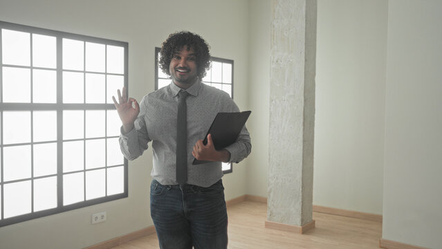 Man holding black folder gestures with open hand and points with other hand in empty building interior, smiling and presenting the space; confidence trust.