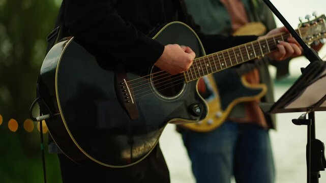 Musicians performing acoustic guitar and bass duet at an outdoor riverside concert during sunset with close-up focus on instruments and live vocal performance