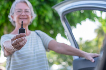Senior adult woman smiling and holding out car key, standing by an open car door, symbolizing...