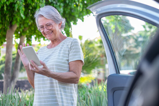 Senior woman smiling and waving during a video call on her digital tablet next to an open car door, enjoying modern communication outdoors