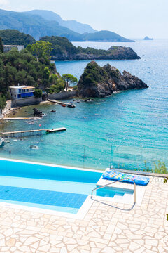 An infinity swimming pool overlooks a turquoise sea bay with a rocky coastline and mountains in Corfu,Greece. A small house and pier sit near the edge of the clear blue water.