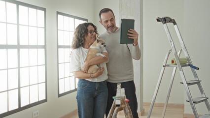 Man and woman, middle aged couple, smile as woman holds small dog and man's hand holds tablet to take a selfie in building  home renovation joy. © Krakenimages.com