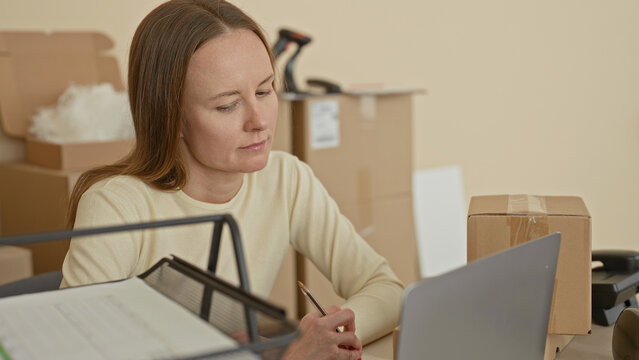 Woman at desk with laptop and parcel, hand holding pencil near keyboard, surrounded by stacked cardboard boxes and barcode scanner in building; pensive small business packing.