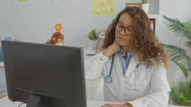 Woman doctor in clinic touches neck in pain while seated at computer with stethoscope and pill bottle visible on desk; tired concern.