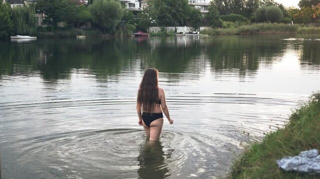 Rear view of a woman in a black swimsuit walking into calm water for an early morning swim in a peaceful natural setting