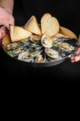 A person holding a pan of cooked mussels in white wine sauce with bread slices, against a dark background. © pavel siamionov