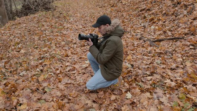 Male photographer in olive parka with fur-trimmed hood kneels on leaf litter in autumn woodland, raises telephoto camera to eye, then lowers it to inspect captured scene under soft overcast light