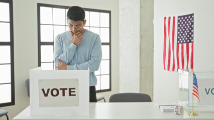 Man touching chin by ballot box in building polling station, wearing glasses and sweater, looking...