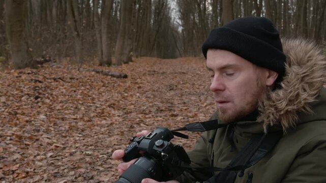 Male photographer in olive parka with fur-trimmed hood kneels on leaf litter in autumn woodland, raises telephoto camera to eye, then lowers it to inspect captured scene under soft overcast light