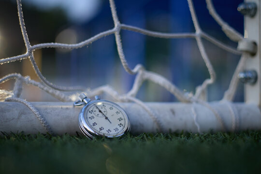 Cinematic close-up of a vintage mechanical stopwatch resting on green grass beside a soccer, with the white net softly blurred in the background. The classic analog dial and precise timing markers