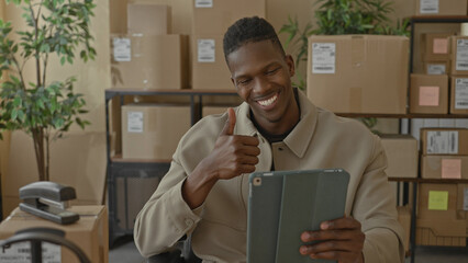 Man in tan jacket holding tablet waves hand and smiles beside stacked parcels on shelving in building  friendly greeting. © Krakenimages.com