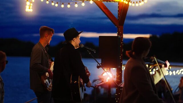 Intimate lakeside evening concert with live acoustic band performing under warm string lights at twilight, creating a relaxed waterfront music atmosphere