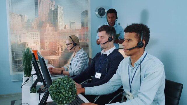 Five call center agents wearing headsets are working collaboratively at desks in a modern office with computers and city views through large windows.