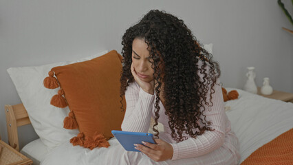 Woman in pink pajamas holding a blue tablet, hand to cheek and furrowed brow while seated on bed in a bedroom with orange pillow and blanket  quiet concern. © Krakenimages.com