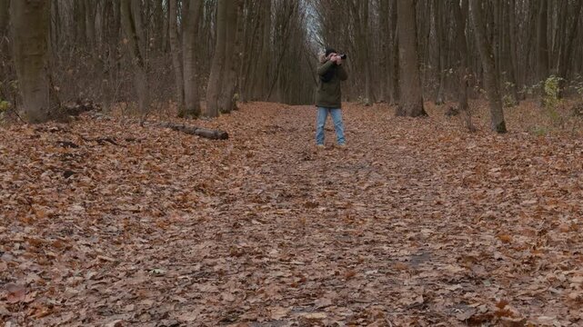 Male photographer in olive parka with fur-trimmed hood kneels on leaf litter in autumn woodland, raises telephoto camera to eye, then lowers it to inspect captured scene under soft overcast light