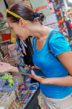 A young female tourist holding a digital tablet browses colorful souvenirs displayed outside a shop while exploring a street market.