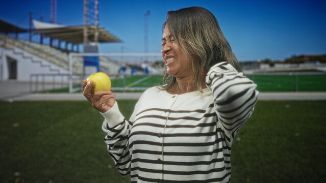 Woman holding lemon, hand on neck and grimacing while inspecting fruit in a stadium building with bleachers and turf visible; disgust.