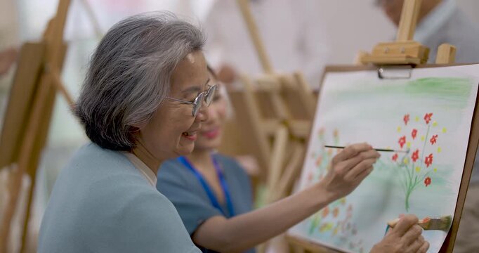 Senior Asian woman paints colorful flowers at easel while female caregiver sits nearby during creative art activity in assisted living center, supporting relaxation, artistic expression and wellbeing.