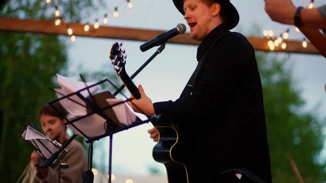 Outdoor evening concert with guitarist and singer performing live music under string lights on a small stage during a relaxed open air gathering