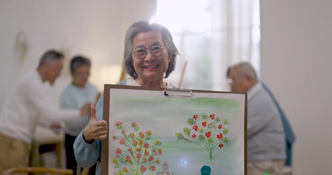 Senior Asian woman presents completed painting and smiles proudly in assisted living community, reflecting creative expression, confidence, emotional wellbeing, relaxation, and active aging.