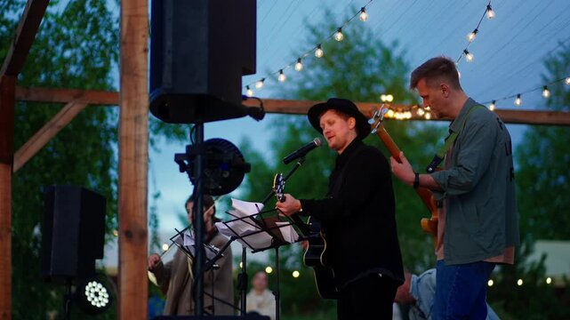 Outdoor evening concert with acoustic band performing live music under string lights on rustic wooden stage surrounded by trees and relaxed audience atmosphere