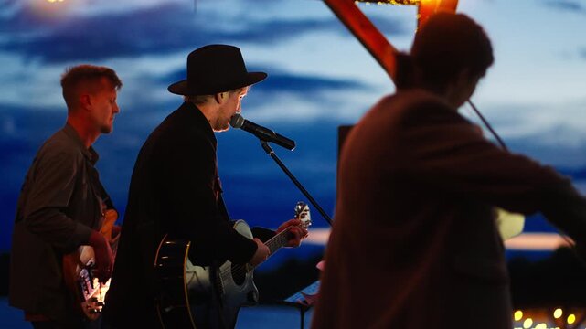 Outdoor lakeside band performing live acoustic music at twilight under string lights with guitarists and singer creating an intimate evening concert atmosphere