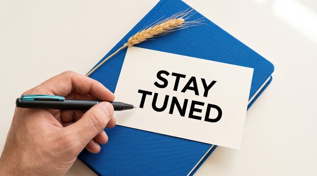Hand writing stay tuned on white paper card placed over blue notebook with a decorative wheat stalk on white surface