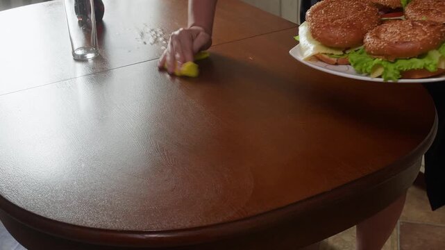 Person wipes down wooden table surface while plate of fresh sandwiches sits nearby, captured with static camera in indoor residential setting with natural lighting.