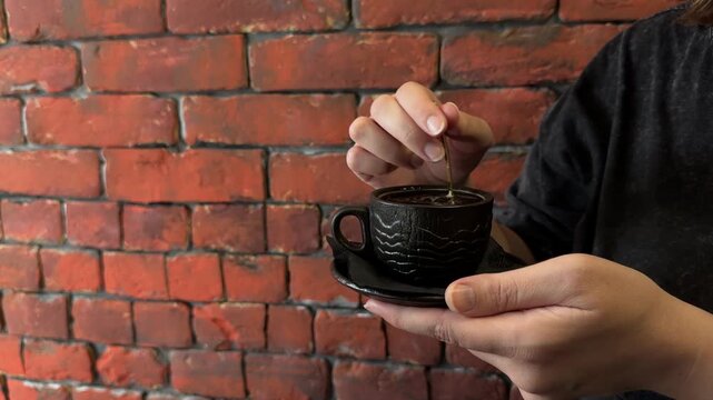 Horizontal video of an unrecognizable person holding a textured black ceramic cup and saucer while stirring hot coffee with a golden teaspoon against a rustic red brick wall with copy space for text