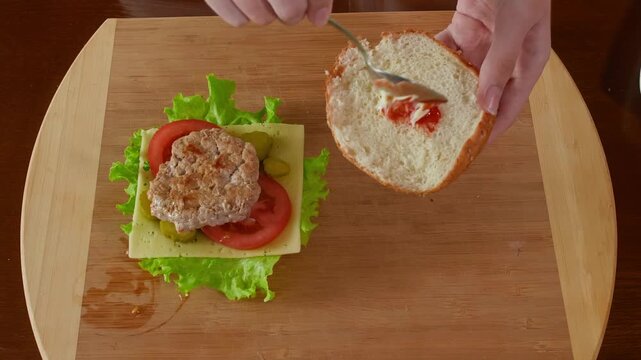 Hands spread red condiment ketchup on bread slice with knife while assembling burger with patty, lettuce, tomato, cheese on wooden cutting board, overhead camera angle.