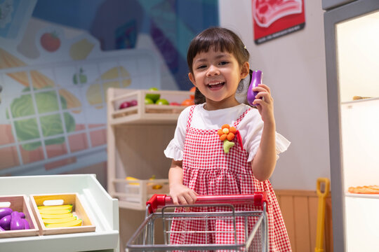 Happy little girl in a red checkered dress shopping for toy vegetables in a play kitchen, smiling with joy and imagination, promoting healthy food choices, creativity, learning, and childhood fun.