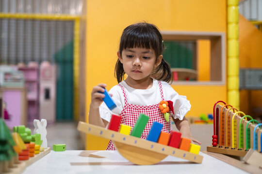 Young girl learning with colorful wooden balance scale and educational toys in a preschool classroom, developing counting, sorting, coordination, concentration, problem solving, and early math skills.