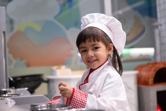 Happy young girl wearing a chef hat and apron, smiling while cooking in a kitchen, representing childhood creativity, playful learning, culinary skills, imagination, family lifestyle, food education.